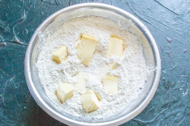 Нарезаем кубиками холодное сливочное масло и добавляем к мучной смеси. Если масло заморожено, натираем его на крупной терке для удобства смешивания.